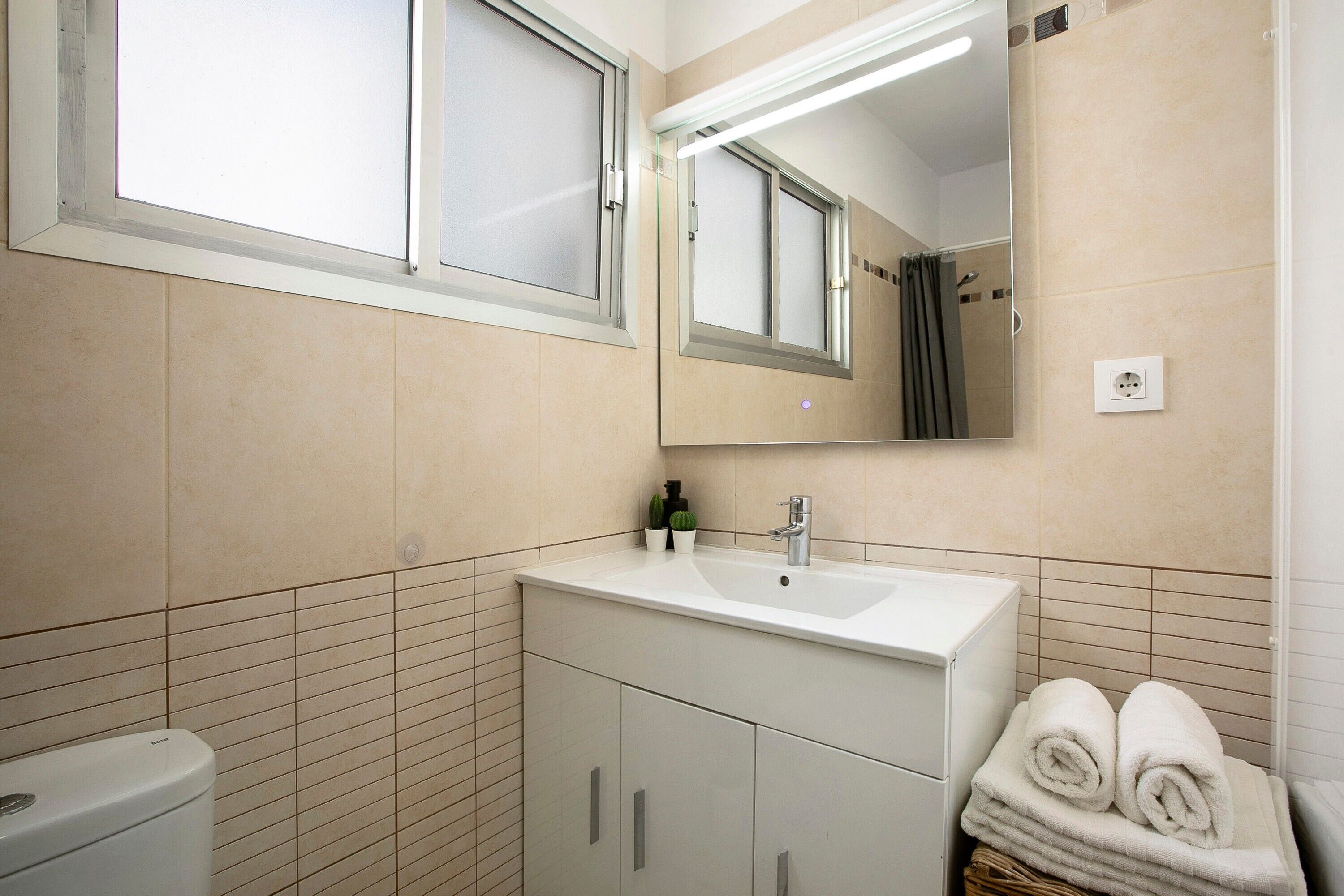 Une salle de bain avec un lavabo blanc, un miroir et des toilettes. Des serviettes sont pliées et empilées dans un panier.