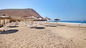 A beach with sun loungers and umbrellas, a clear sky, and a mountain in the background.