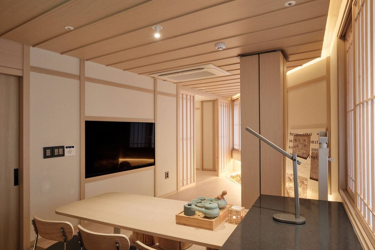 Un interior moderno con techo de madera, televisor de pantalla plana, mesa de comedor con sillas y una zona de cocina con estufa y tetera.