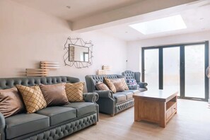 A spacious living room with a large window, a sofa set, a wooden coffee table, and a wall-mounted mirror. - Lush & Spacious 3BD House - East Brighton (Brighton)