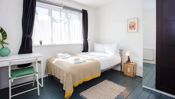 A bedroom with a bed, a desk, a chair, a small table with a lamp, a potted plant, and a window with curtains. - Stylish 4BD House With Private Garden - Stratford (London)