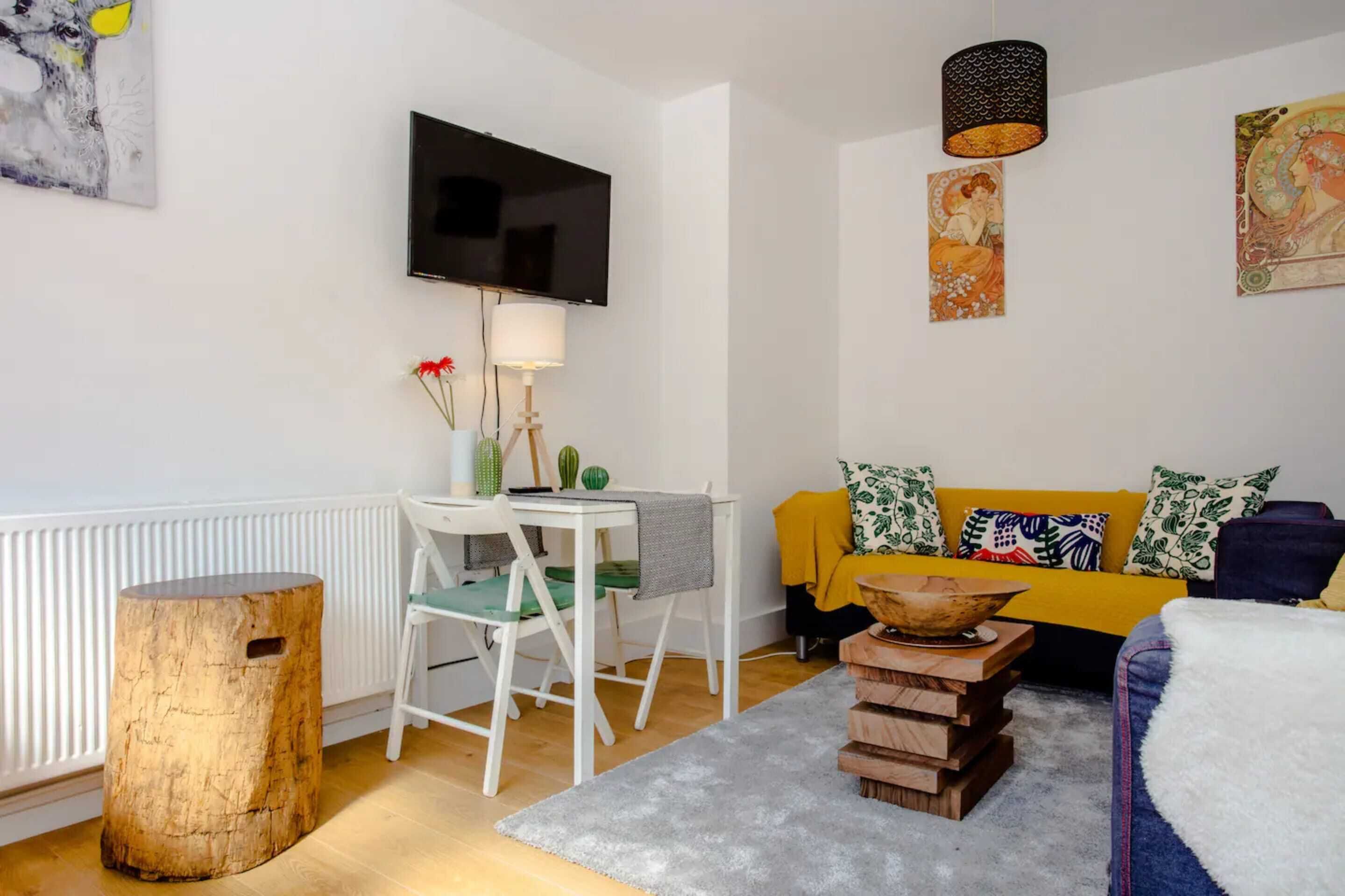 A modern living room with a flat-screen TV, a wooden side table, a sofa with patterned cushions, and a decorative lamp.
