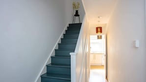 A staircase with white railings and a dark-colored handrail, leading to a bright room with a door and a small table with a plant. - Stylish 4BD House With Private Garden - Stratford (London)