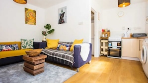 A modern living room with a sofa, a coffee table, and a kitchen area with a washing machine. - Stylish 4BD House With Private Garden - Stratford (London)