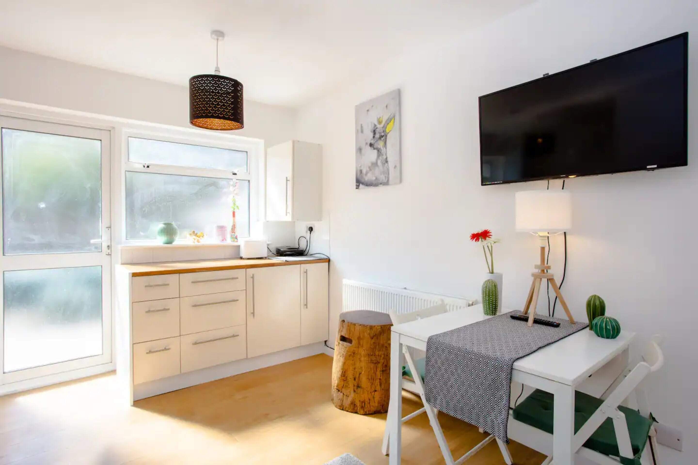 A modern kitchen with white cabinets, a wooden floor, a dining table with a chair, a TV mounted on the wall, and a window with frosted glass.