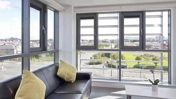 Una habitación moderna con sofá de cuero negro, ventanas amplias que ofrecen vistas a la ciudad y una pequeña mesa con una planta en maceta.