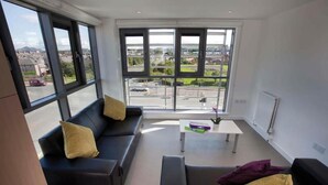 Una sala de estar moderna con un sofá de cuero negro, una mesa de centro blanca y amplios ventanales con vistas a un paisaje urbano.