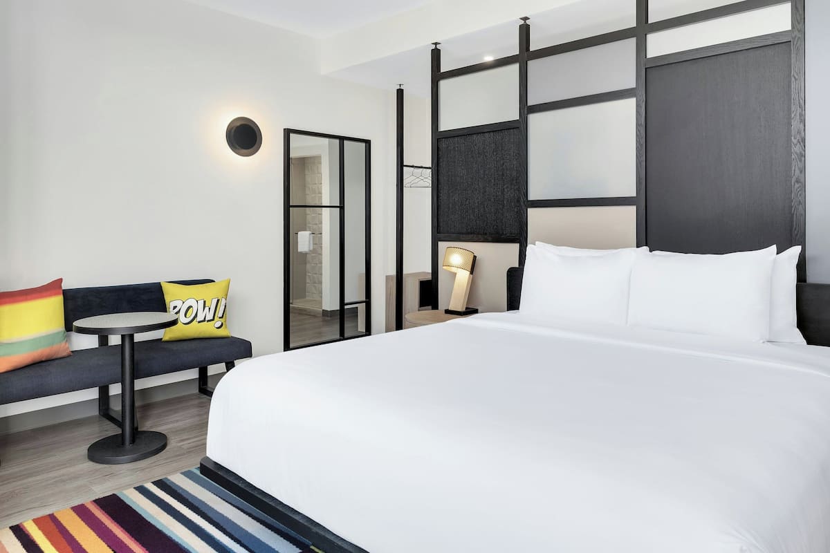 A hotel room with a large bed, a small round table, a sofa with colorful cushions, a mirror, and a wall-mounted light fixture.