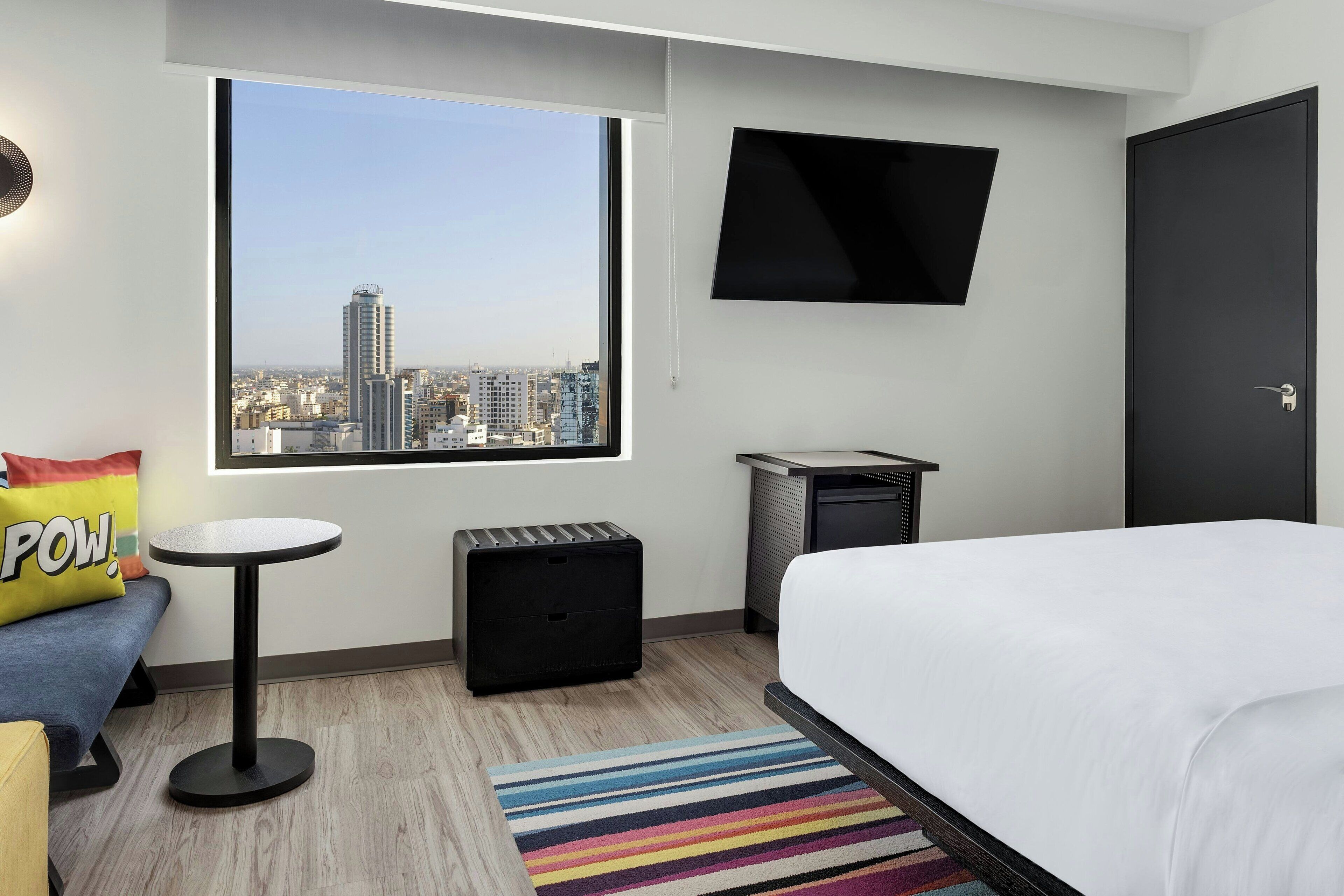 Une chambre d’hôtel moderne dotée d’une grande fenêtre, d’un lit, d’une petite table ronde, d’un téléviseur fixé au mur et offrant une vue sur la ville.