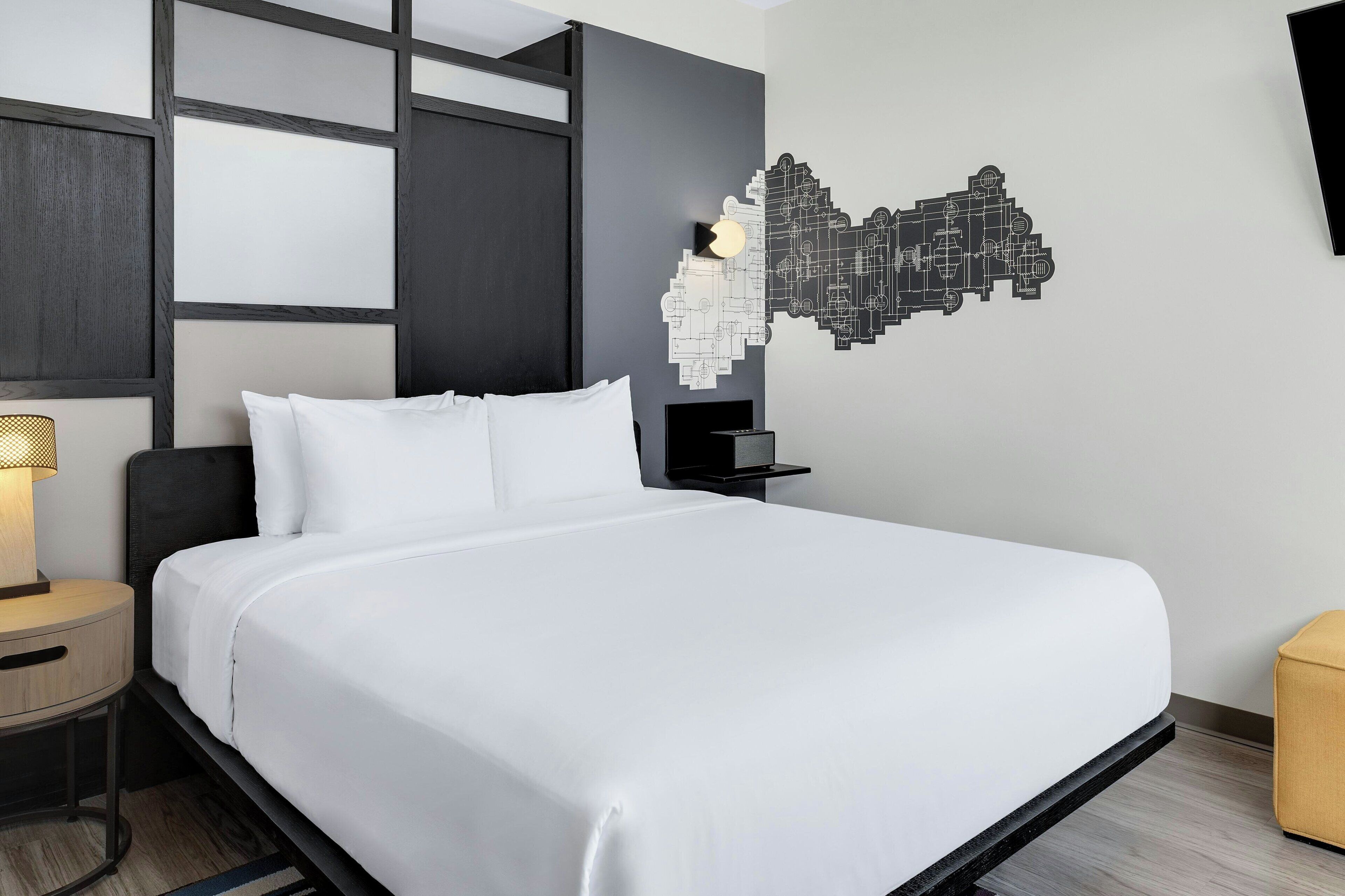 Habitación de hotel moderna con una cama grande, una mesita de noche con lámpara, un televisor montado en la pared y una decoración geométrica única en la pared.