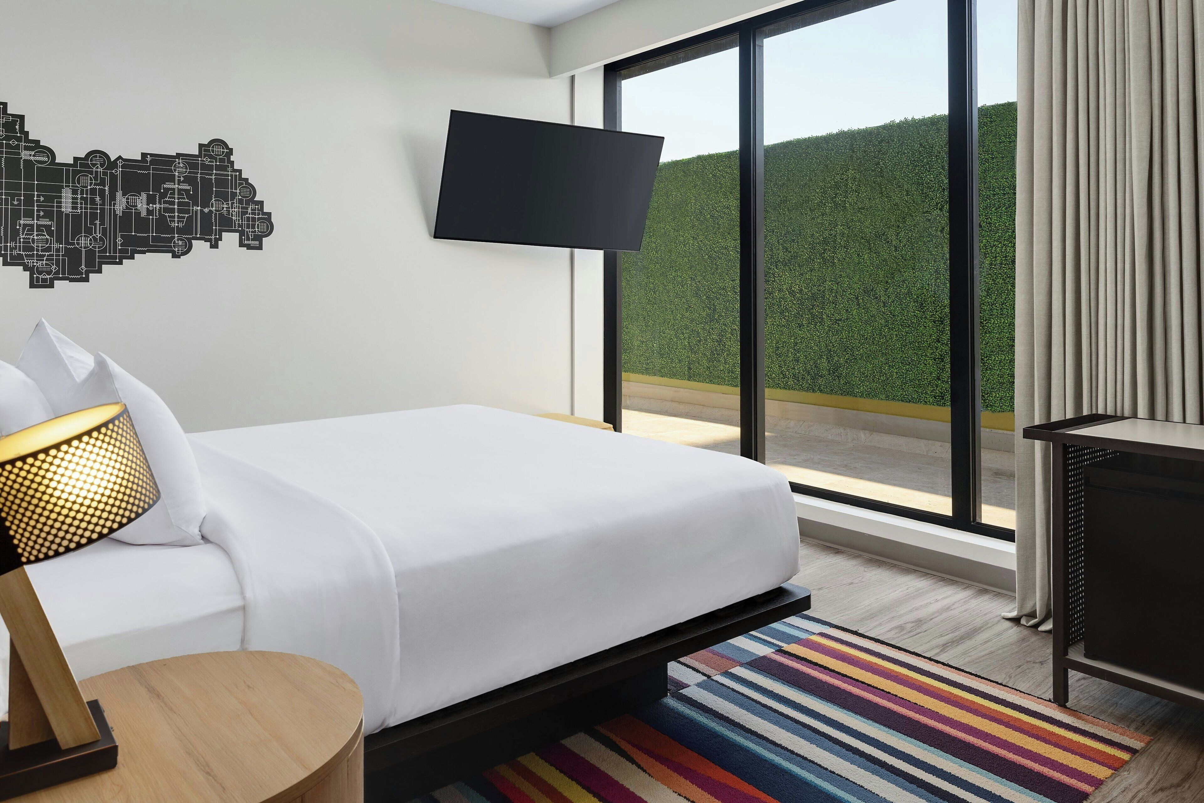 Een moderne hotelkamer met een groot bed, een flatscreen televisie aan de wand, een ronde houten bijzettafel en een kleurrijk gestreept tapijt.
