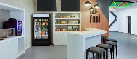 A modern bar area with a white counter, black stools, and a 'Fuel' sign. There is a refrigerator stocked with beverages and snacks, and a microwave on the counter.