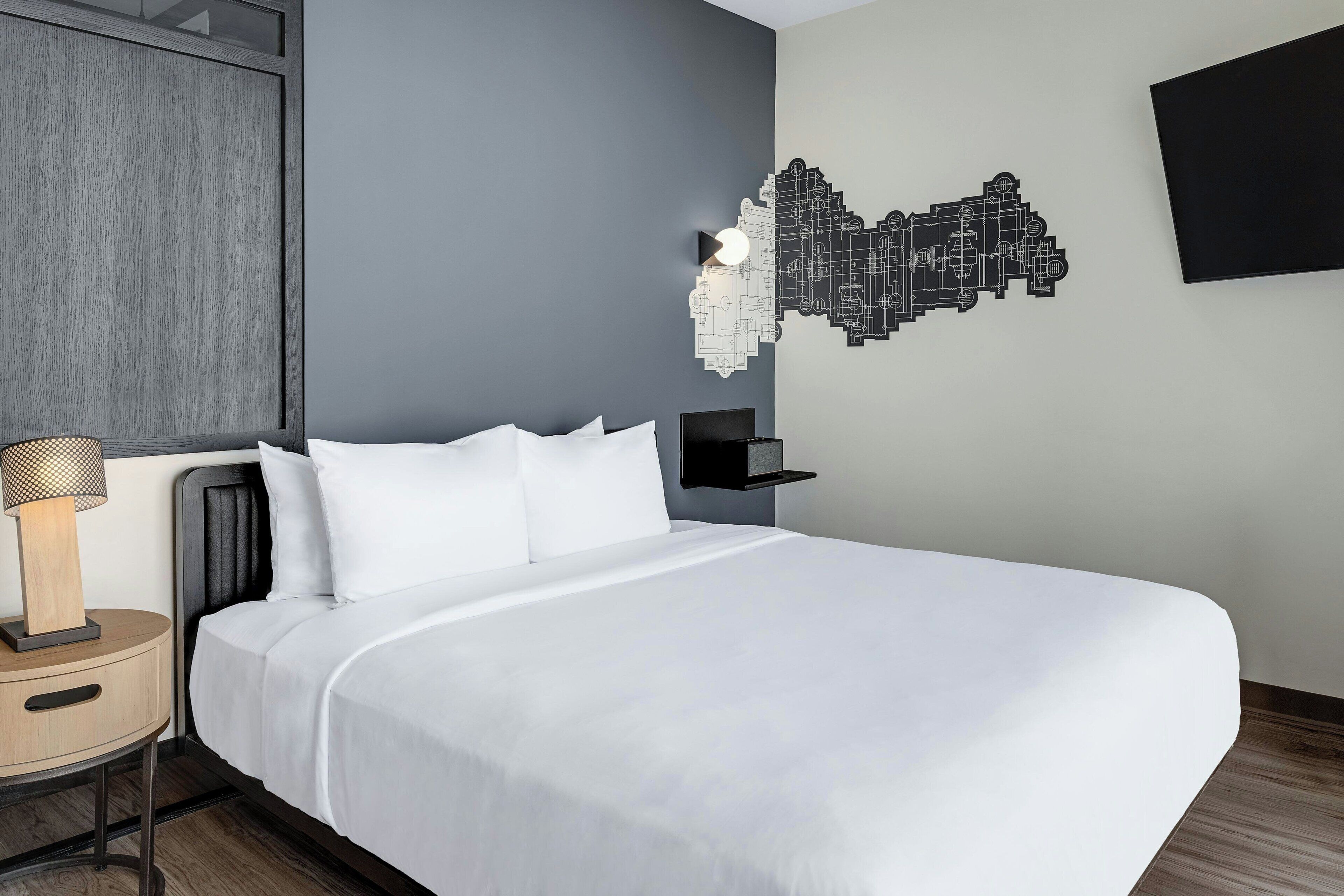 A modern hotel room with a large bed, a bedside table with a lamp, a wall-mounted TV, and a decorative wall piece.