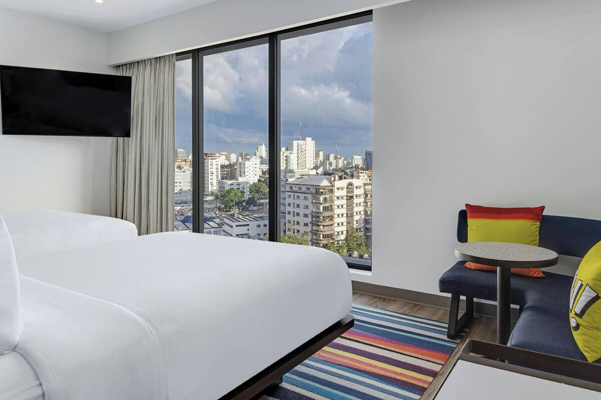 A hotel room with a large window, a bed, a sofa, and a small table.