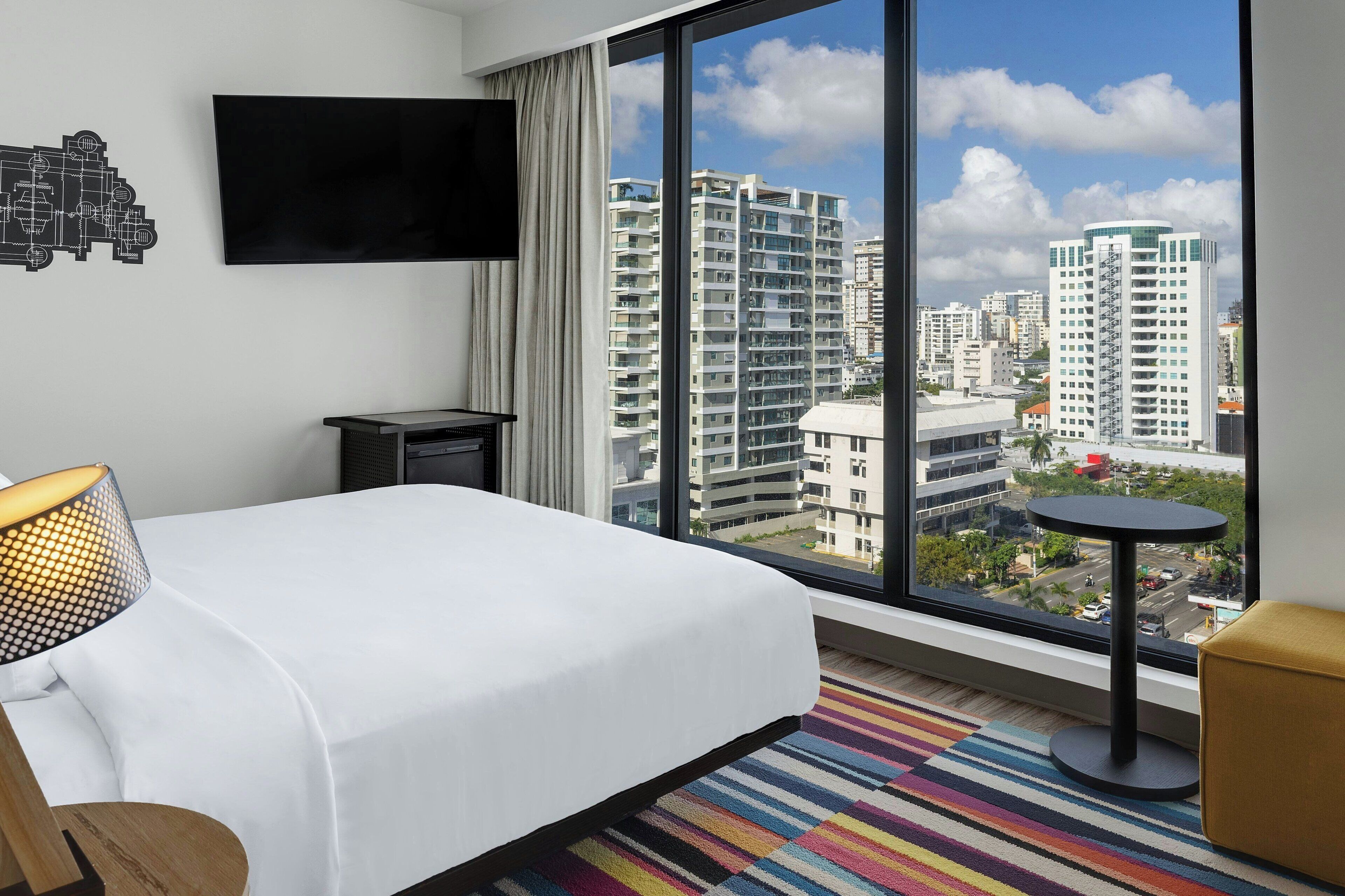 A hotel room with a large window, a bed, a TV, a small table, and a colorful striped rug.