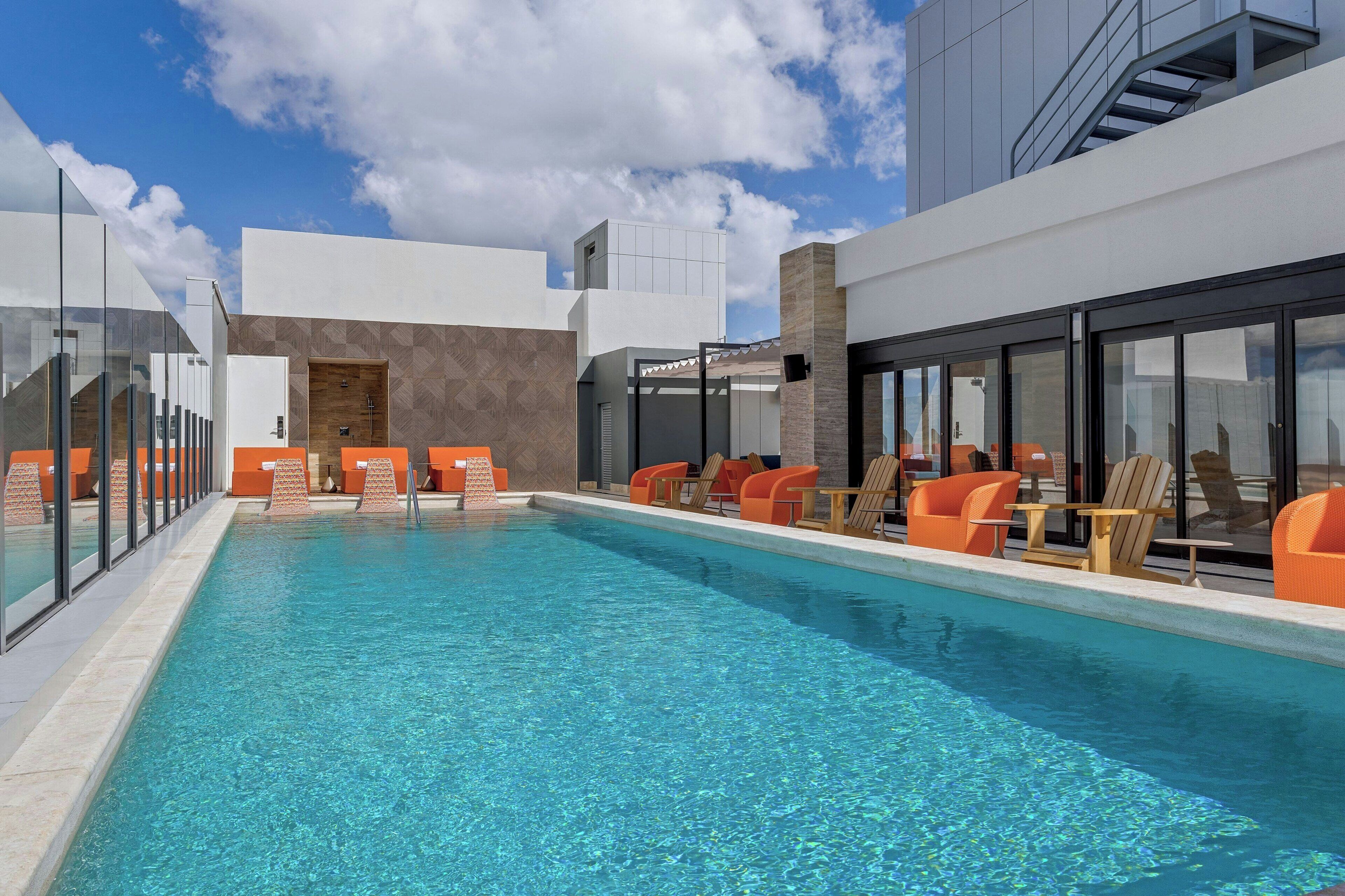 A modern outdoor pool with clear blue water, surrounded by orange chairs and tables, and large glass windows.