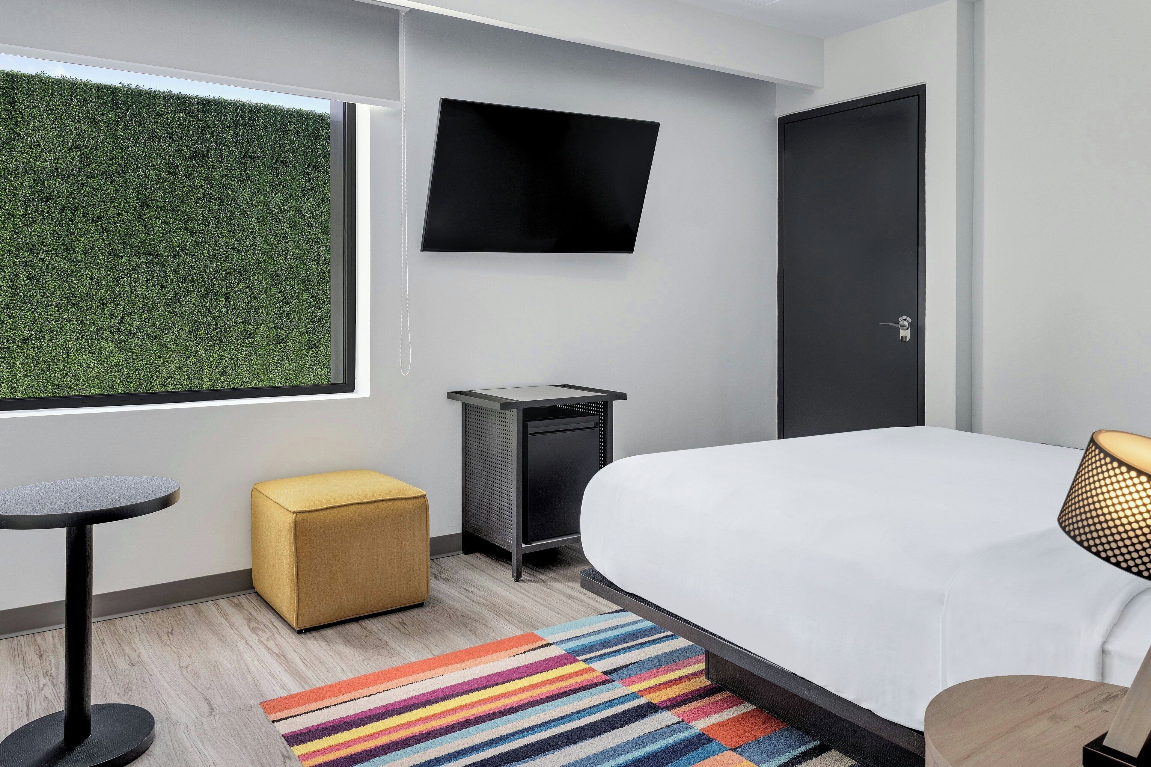 Une chambre d’hôtel moderne dotée d’un grand lit, d’un téléviseur à écran plat fixé au mur, d’un petit bureau et d’une fenêtre donnant sur une haie verdoyante.