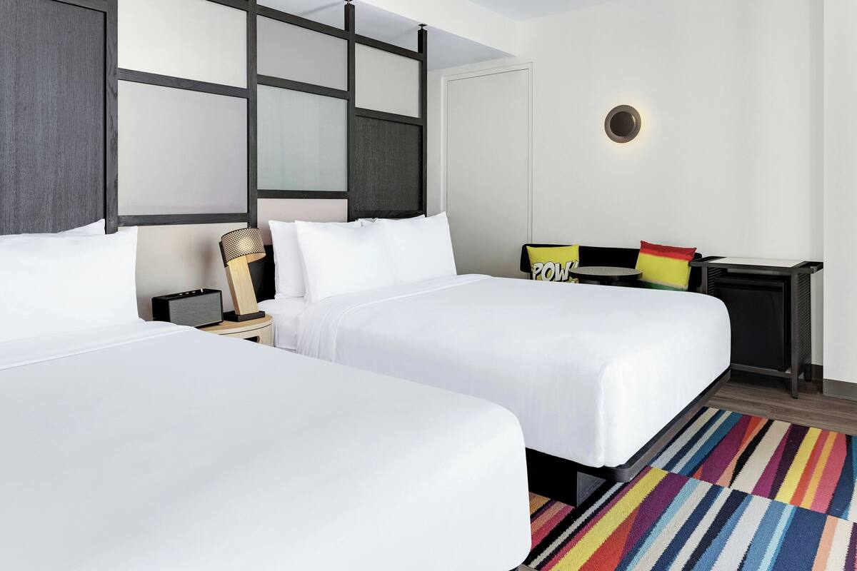 A hotel room with two beds, a small table with a lamp, a colorful striped rug, and a wall-mounted light fixture.