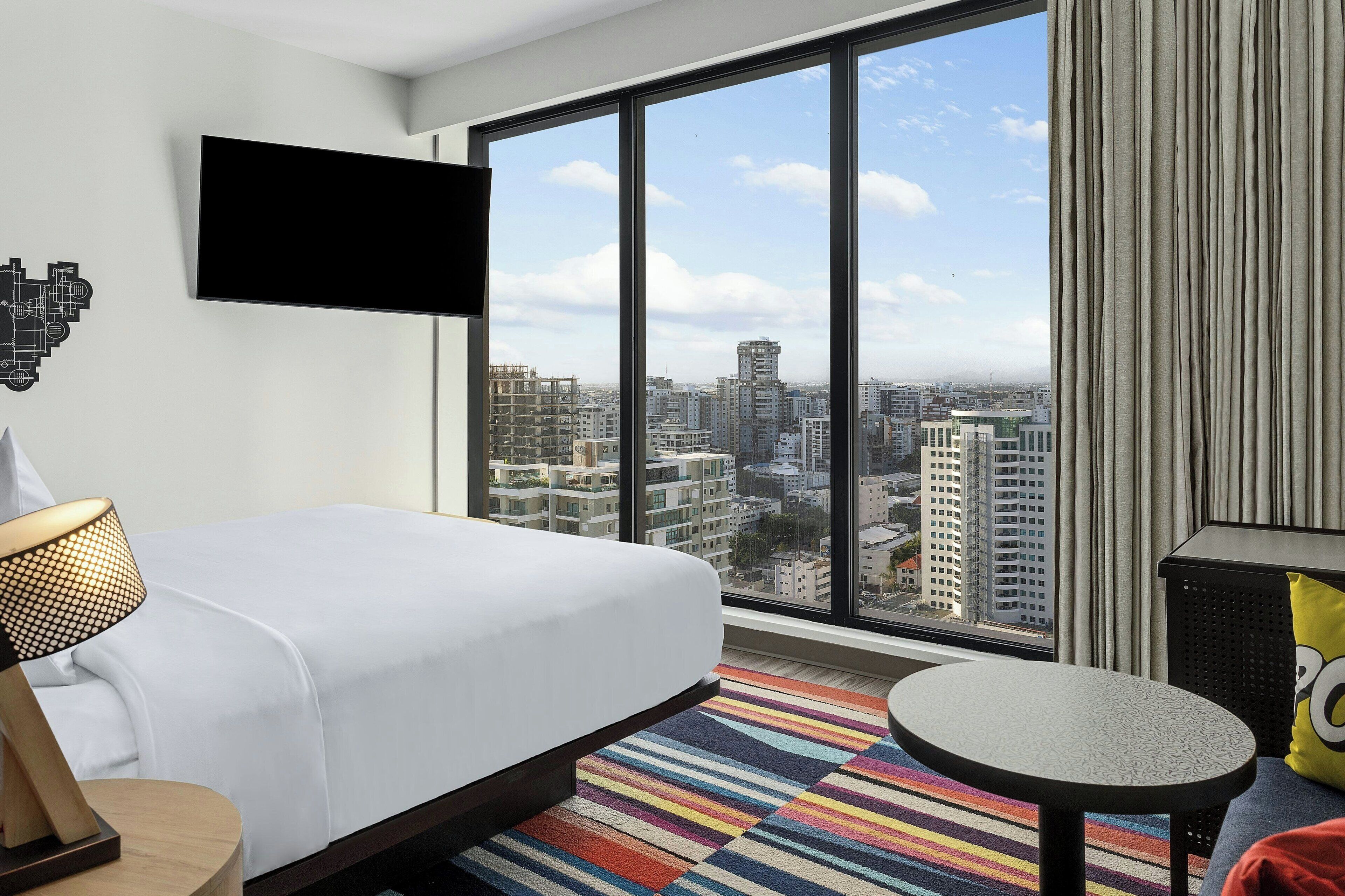 A hotel room with a large bed, a TV mounted on the wall, a small round table, and a colorful striped rug.