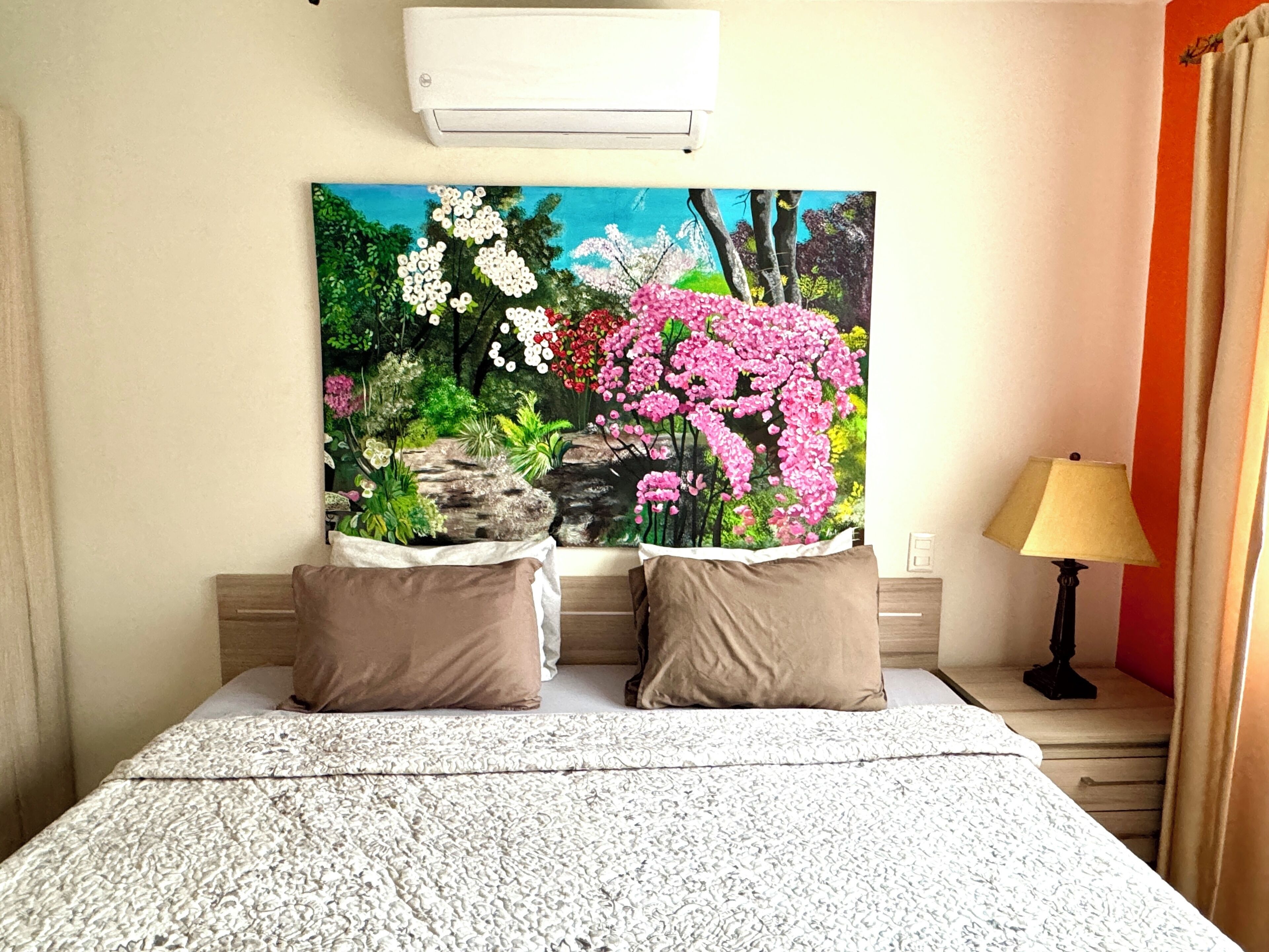 A bedroom with a bed, two pillows, a nightstand with a lamp, and a large floral painting on the wall.