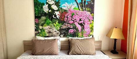 A bedroom with a bed, two pillows, a nightstand with a lamp, and a large floral painting on the wall.