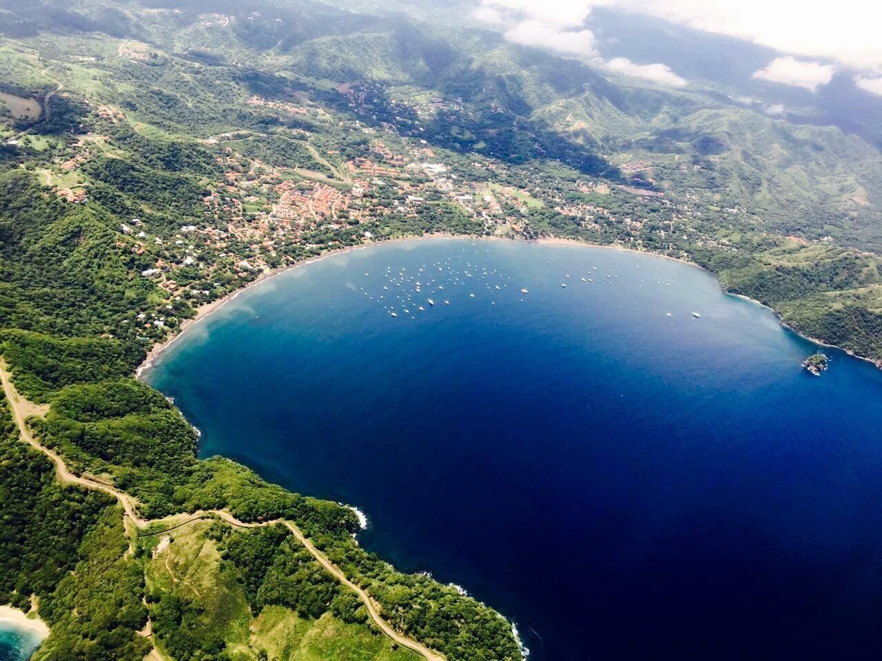An aerial view of a coastal area with a large bay, surrounded by dense greenery and a small town.