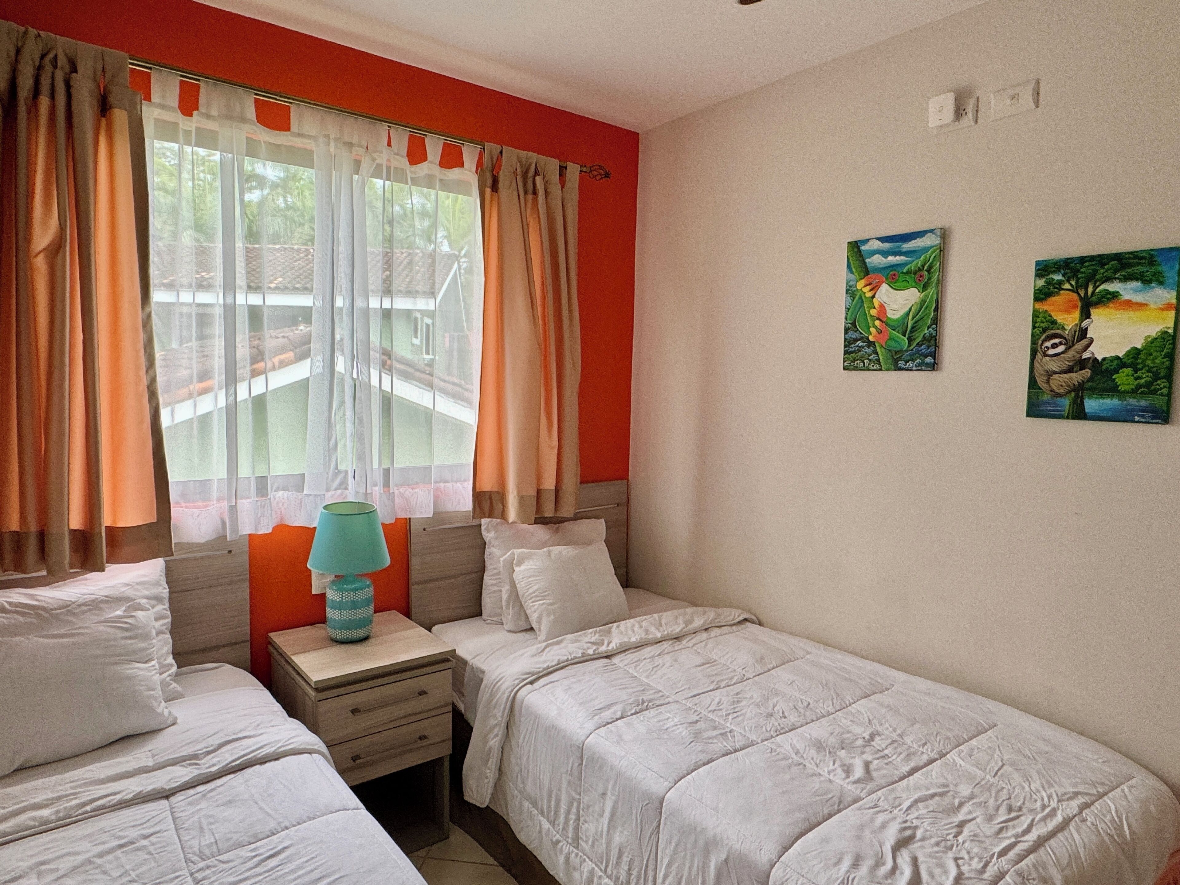 A room with two single beds, a nightstand with a lamp, and a window with curtains. There are two paintings on the wall.