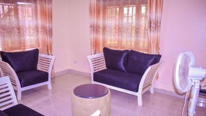 A room with two white armchairs with dark cushions, a round ottoman, and a standing fan. - The Spot Hotel (Mbale)