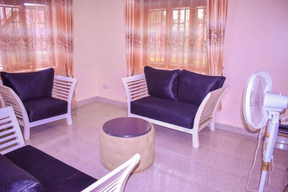 A room with two white armchairs with dark cushions, a round ottoman, and a standing fan.
