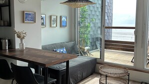 A modern living space with a wooden dining table, black chairs, a grey sofa, and a large window offering a view of the beach. - Vue sur Mer, Appartement Rénové au Coeur de la Station Balnéaire (Agon-Coutainville)