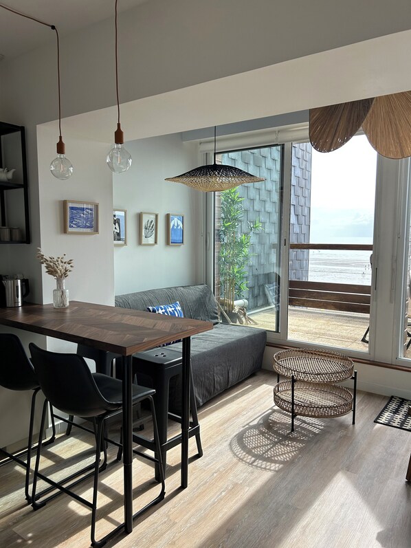Ein modernes Wohnzimmer mit einem Holzespeisetisch, schwarzen Stühlen, einem grauen Sofa und einem großen Fenster mit Blick auf den Strand.