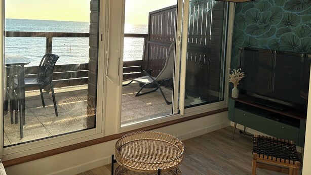 A room with a large sliding glass door leading to a balcony with a view of the sea. There is a wicker chair and a small table on the balcony, and a television on a stand in the room. - Vue sur Mer, Appartement Rénové au Coeur de la Station Balnéaire (Agon-Coutainville)