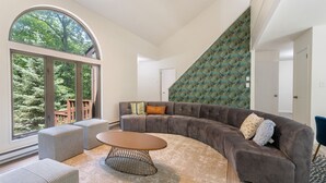 Een moderne woonkamer met een gebogen grijze bank, een ronde salontafel en een groot raam met uitzicht op bomen.