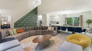 Een moderne woonkamer met een hoekbank, een ronde salontafel en een gele fauteuil. Een trap met een patroonwand, een eethoek met blauwe stoelen en een keuken op de achtergrond.
