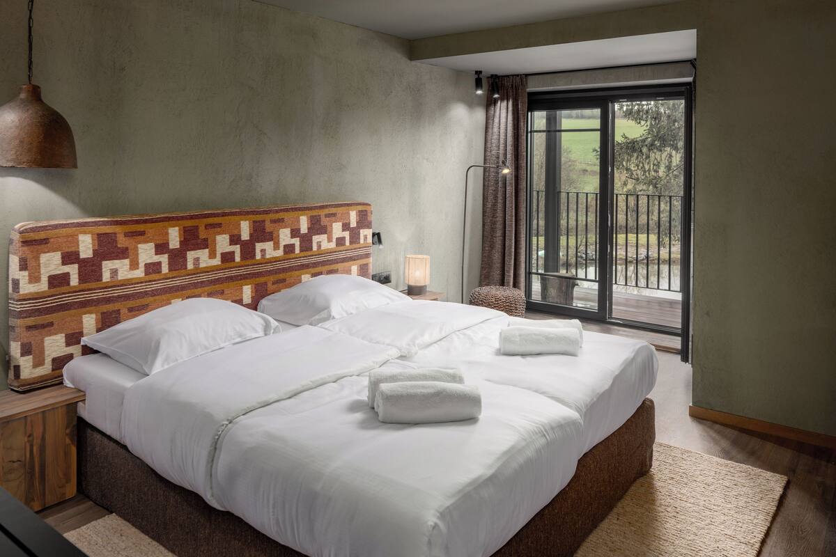 Een groot bed met witte lakens en twee handdoeken, een hoofdbord met een patroon, een nachtkastje met een lamp, en uitzicht op de omgeving door een schuifdeur.