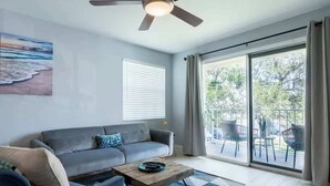 Uma sala de estar com sofá, mesa de centro, ventilador de teto, um quadro e uma porta de vidro deslizante que dá para uma área externa com cadeiras.