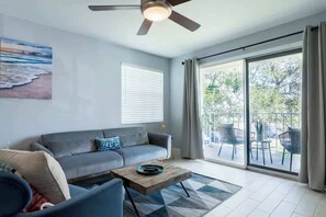 A living room with a sofa, a coffee table, a ceiling fan, a painting, and a sliding glass door leading to an outdoor area with chairs.