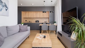 Una sala moderna con un sofá gris, una mesa de centro de madera y un mueble para la televisión. Se observa una zona de cocina con un armario de madera y una mesa de comedor con sillas.