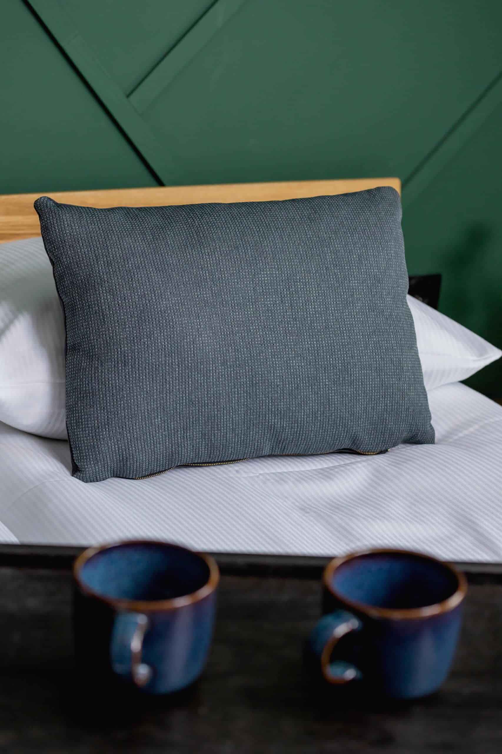 Un lit avec un oreiller gris et deux tasses bleues sur une table en bois.
