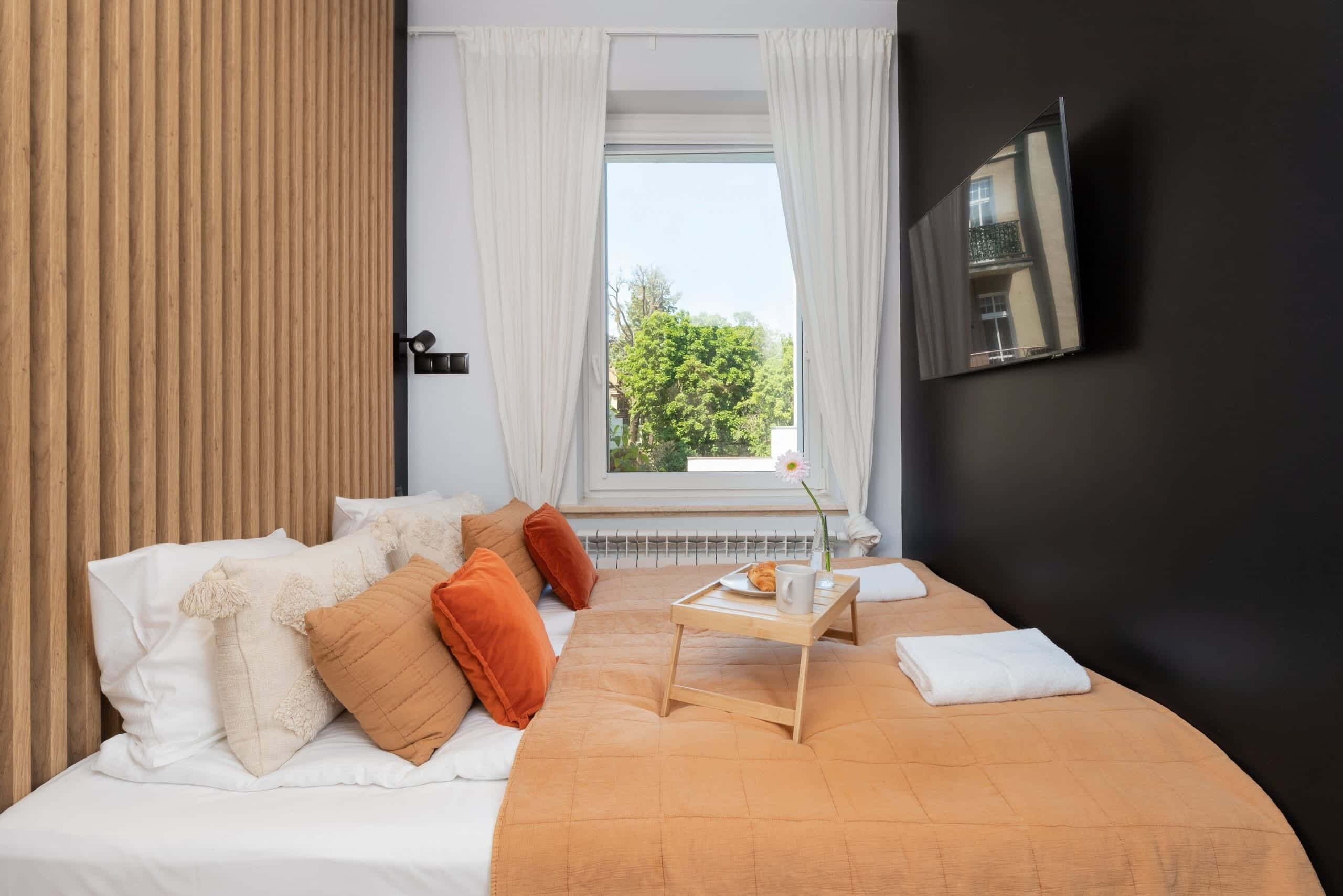 Et pænt arrangeret soveværelse med seng, puder, et natbord med bakke, et vindue med gardiner og et TV monteret på væggen.