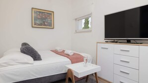 Un dormitorio con cama, televisión, mesita, armario y ventana.