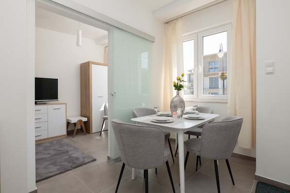 A modern dining area with a white table, four grey chairs, a vase with flowers, and a window with curtains.