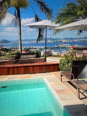 A pool with a view of a city and the sea, a bench, and umbrellas.