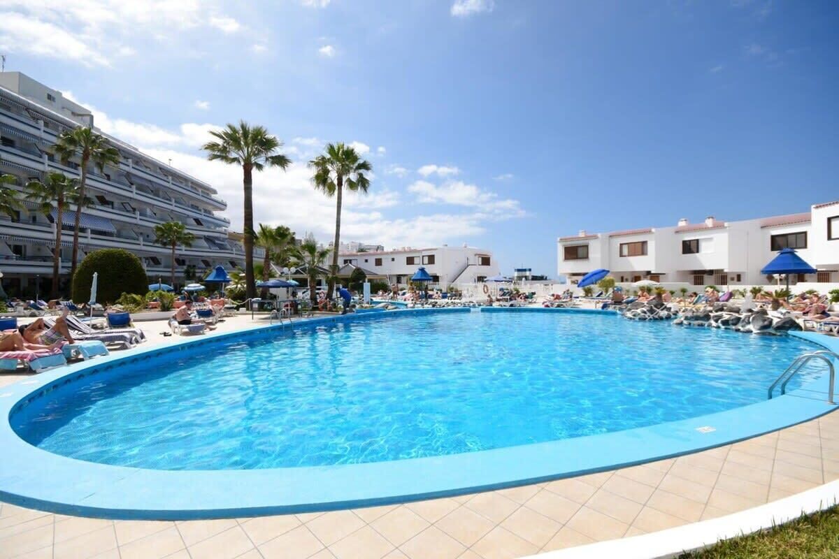 Une piscine entourée de palmiers et d’immeubles, avec des chaises longues et des parasols.