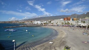 A coastal resort with a sandy beach, a marina with boats, and a backdrop of mountains.