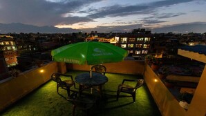 A rooftop terrace with a green umbrella, chairs, and a view of a cityscape and mountains at dusk. - MeroStay 190 HOMESTAY (Sainbu)