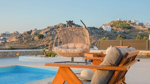 A poolside area with a wicker chair and wooden lounge chairs, overlooking a rocky hillside with buildings. - Eden's Mykonos - Villa Opal (Mykonos)