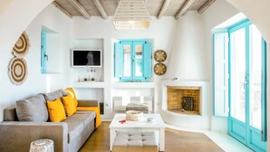 A cozy living room with a sofa, coffee table, and fireplace. There is a television mounted on the wall and a window with blue shutters. - Eden's Mykonos - Villa Opal (Mykonos)