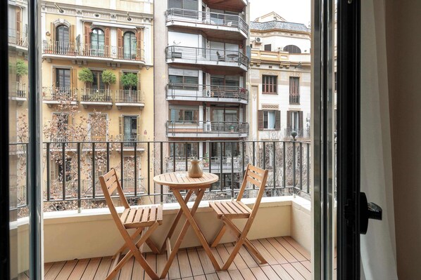 A balcony with a table and two chairs, overlooking a cityscape with multi-story buildings.