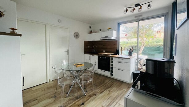 A modern kitchen with a glass dining table, white cabinetry, and a large window offering a view of the outdoors. - Apartment for 4 in Argelès-sur-Mer with garden and garage (Argelès-sur-Mer)